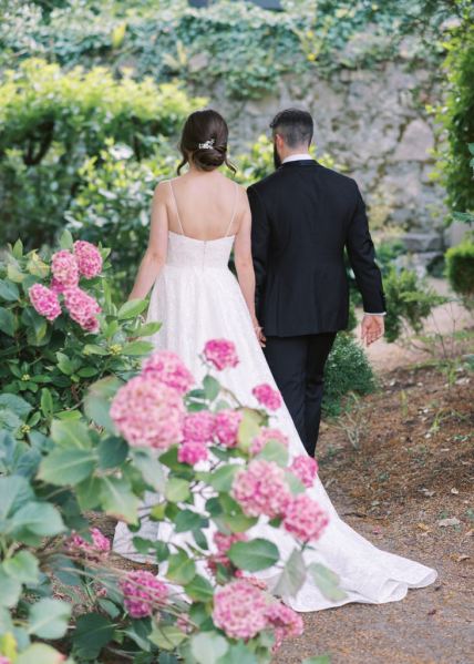 Bride and groom walk in garden hand in hand pink flowers back of dress