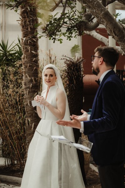 Bride holding paper vows and drink looking at groom giving speech