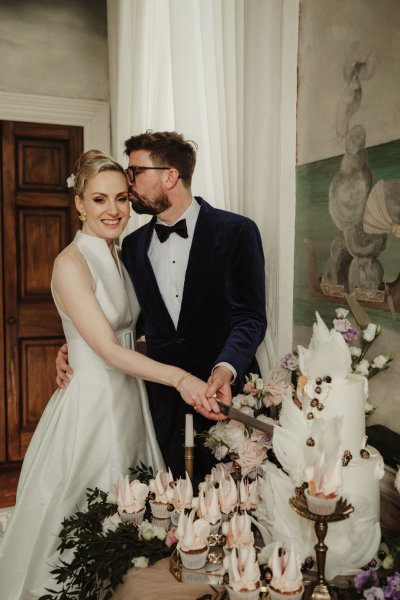 Groom kisses bride on cheek whilst cutting the cake