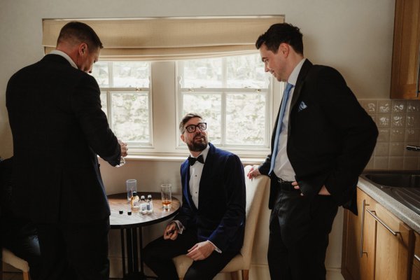 Groom and groomsmen sitting at window drinking whiskey