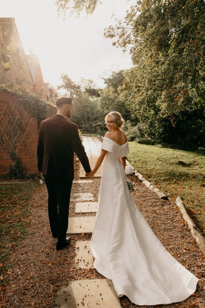 Bride and groom walk along pathway in forest/park holding hands sun is shining over the shoulder