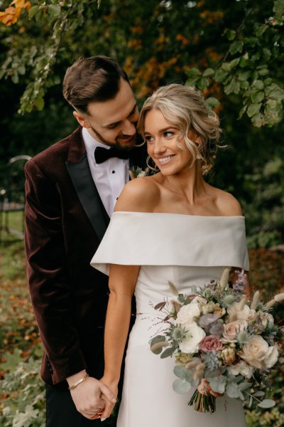 Bride and groom couple surrounded by trees in park smiling whispering in ear