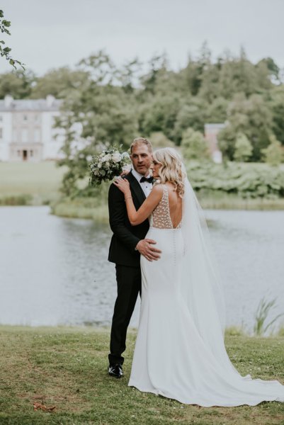 Manor house wedding venue in background as bride and groom embrace on grass lake