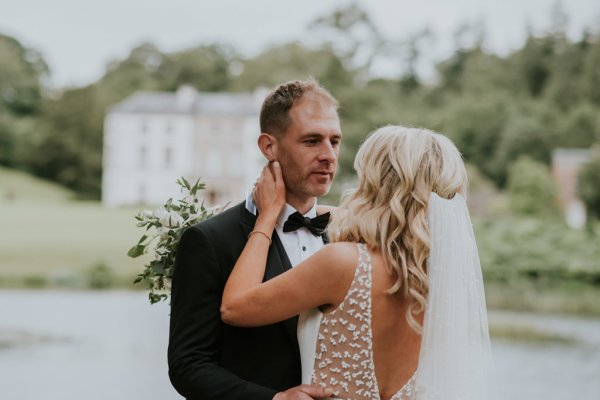 Manor house wedding venue in background as bride and groom embrace on grass lake