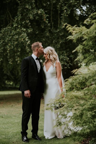 Bride and groom standing on grass in forest kiss on forehead