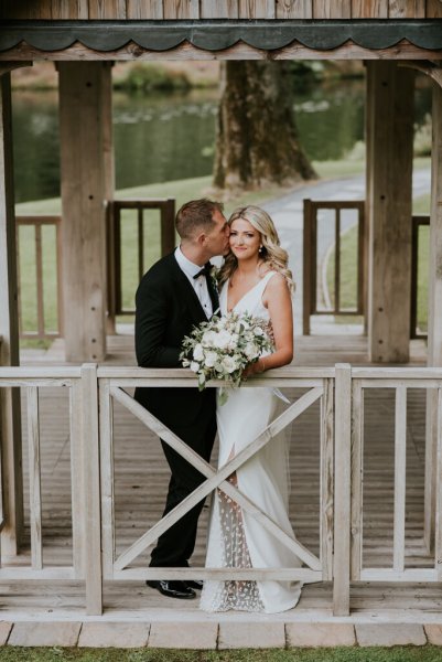 Bride and groom stand on boardwalk above lake