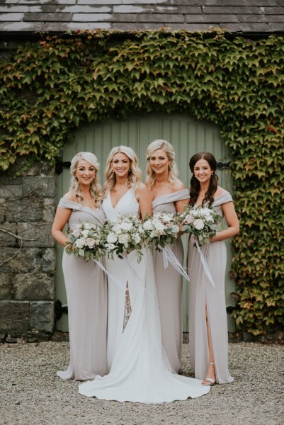 Bride and bridesmaids standing in front of green gate holding bouquet/flowers