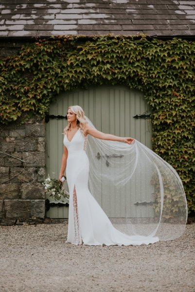 Bride shows of bridal heels/shoes on farm in front of green gate showing off veil detail