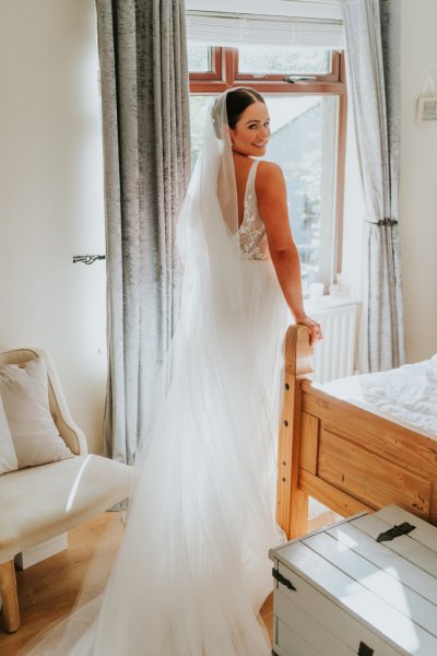 Bride looks over her shoulder back of dress detail and veil standing at window