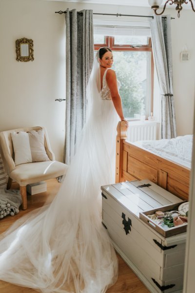 Bride looks over her shoulder back of dress detail and veil standing at window