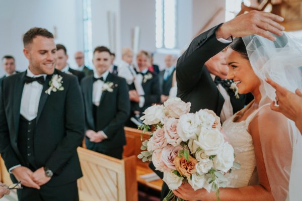 Father takes veil off brides head surrounded by guests holding bouquet of flowers/roses