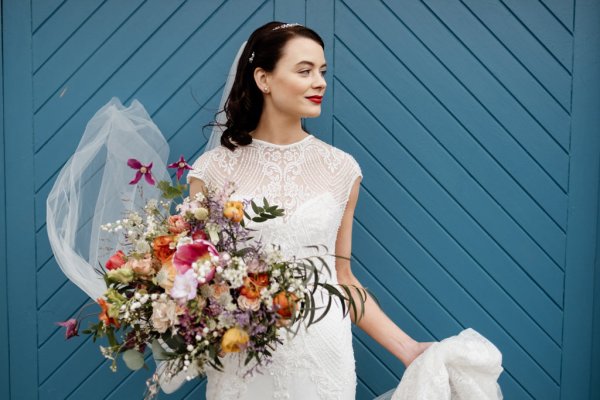 Veil blowing in the wind bride standing in front of blue gate door holding bouquet of flowers