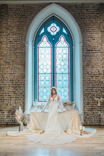 Bride faces camera stands beside dining room table in white window detail bricked wall