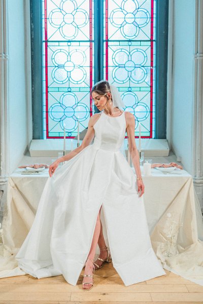 Sleeveless dress bride standing against white dining room table bricked wall showing off dress