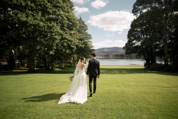 Bride and groom from behind hand in hand standing on grass in front of lake