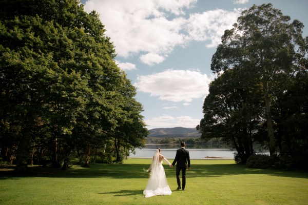 Bride and groom from behind hand in hand standing on grass in front of lake