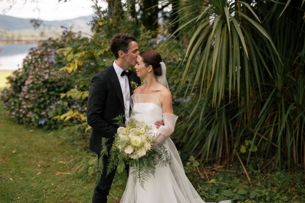 Bride and groom kiss on the forehead in front of trees in garden