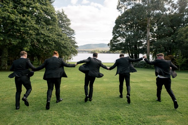 Groom and groomsmen running on grass towards lake