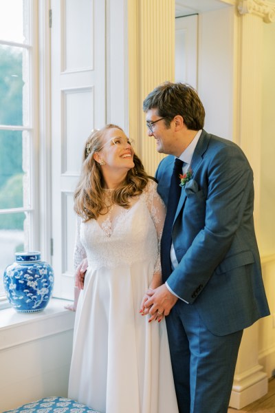 Bride and groom hold hands at window smiling looking at each other