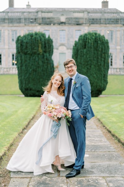 bride and groom stand on pathway in garden holding flowers