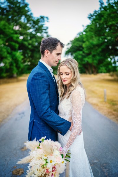 Bride looks down as she and her groom stand in garden holding bouquet of flowers a