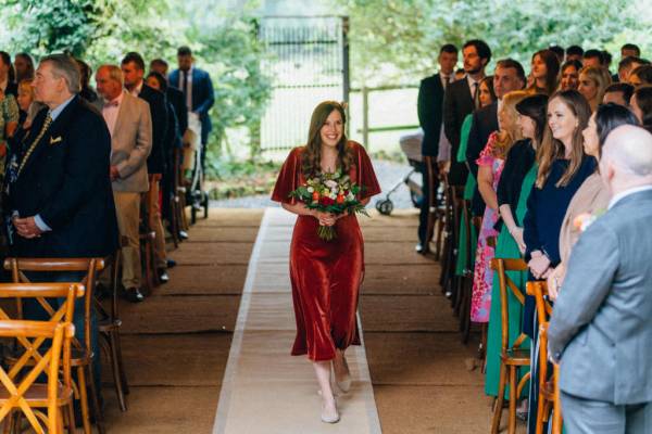 Bridesmaid in satin red dress walks down the aisle carrying flowers