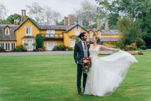 Bride shows off dress groom stands on grass outside wedding venue