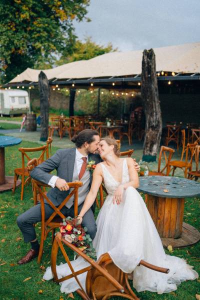 Bride and groom sit on wooden chairs outside in garden area