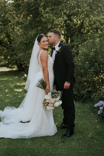 Exterior shot of bride and groom in garden kiss on the cheek