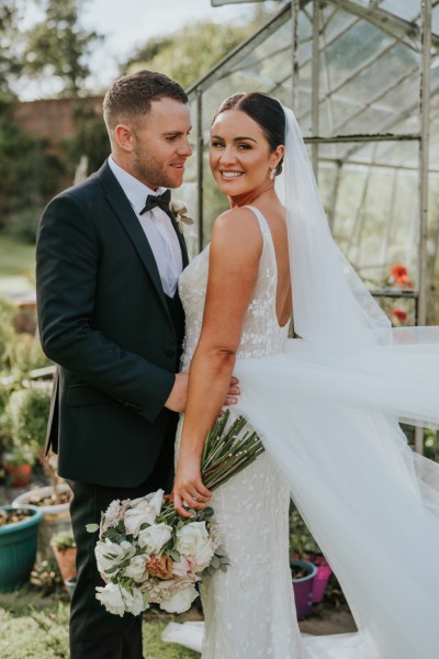 Groom looks at bride arms around waist outside greenhouse flowers bouquet