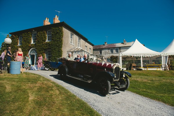 Groom and groomsmen drive the wedding car wedding venue in background with tent/marquee