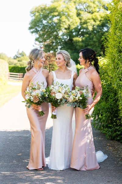 Bride and bridesmaids stand in garden smiling holding bouquet of flowers