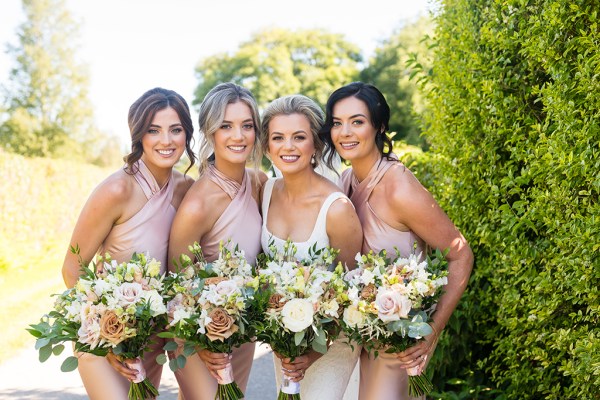 Bride and bridesmaids stand in garden smiling holding bouquet of flowers