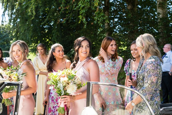 Bridesmaids stand beside wedding car smiling bouquet flowers