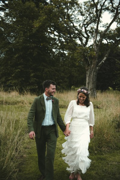 Bride and groom walk hand in hand on grass in forest park setting