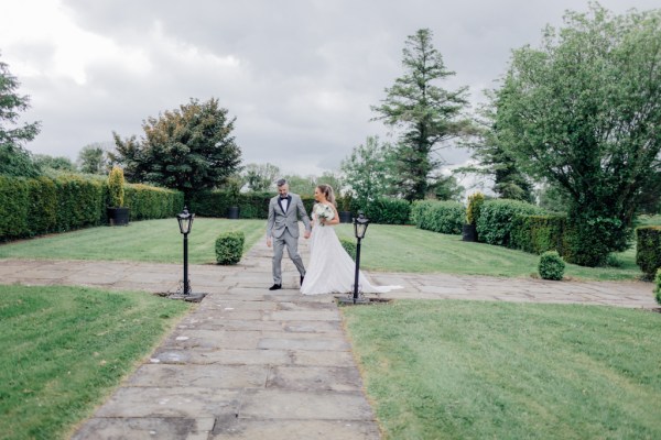 Bride and groom walk outside on the pathway holding bouquet of flowers lanterns on grass