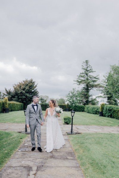 Bride and groom walk outside on the pathway holding bouquet of flowers