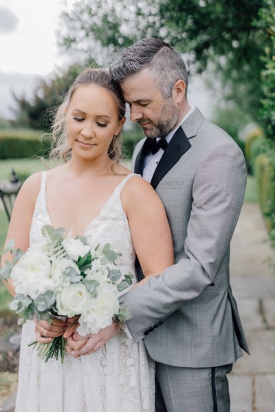 Bride and groom pose outside on the pathway holding bouquet of flowers