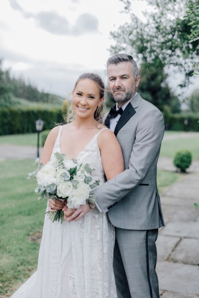 Bride and groom pose outside on the pathway holding bouquet of flowers