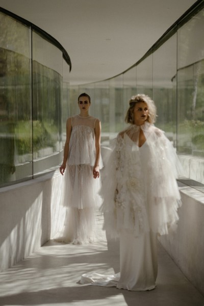 Mid close up of two brides pose for camera wearing white bridal gowns/dresses garden in view behind