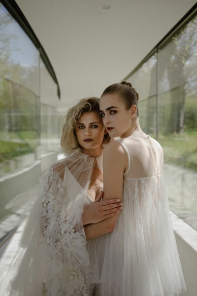 Mid close up of two brides pose for camera wearing white bridal gowns/dresses garden in view behind