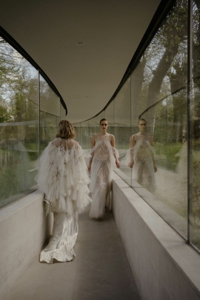 Two models ruffle long bridal white gown dresses window reflection garden in view