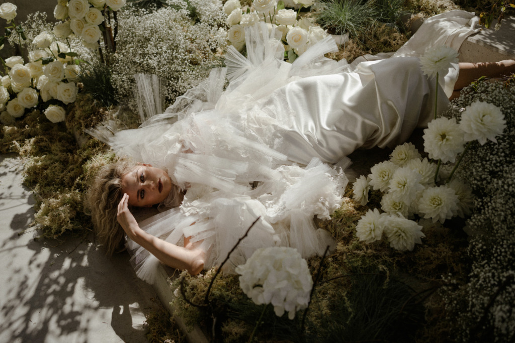 Bride lying down on bed of flowers/white roses poses silky bridal gown dress on