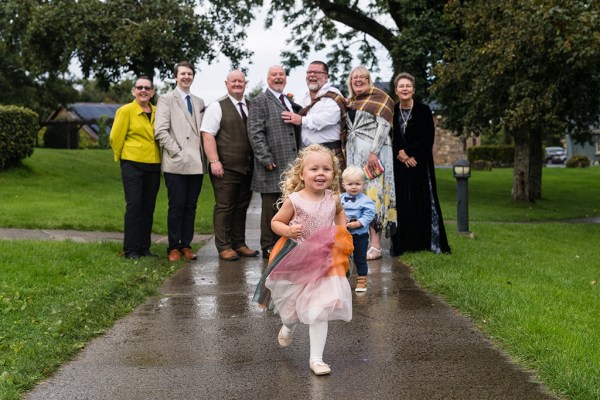 Little girl runs boy in background with grooms and family members in park garden setting