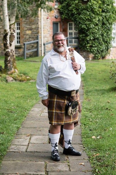 Groom in kilt stands on pathway in garden grass setting