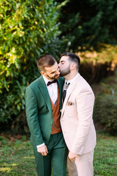 Groom kisses groom on cheek smiling hugging in garden