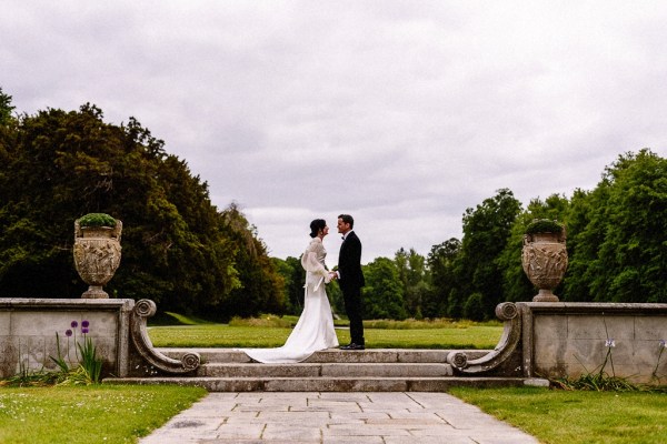 Bride approaches groom in garden grass in background they embrace on steps hug