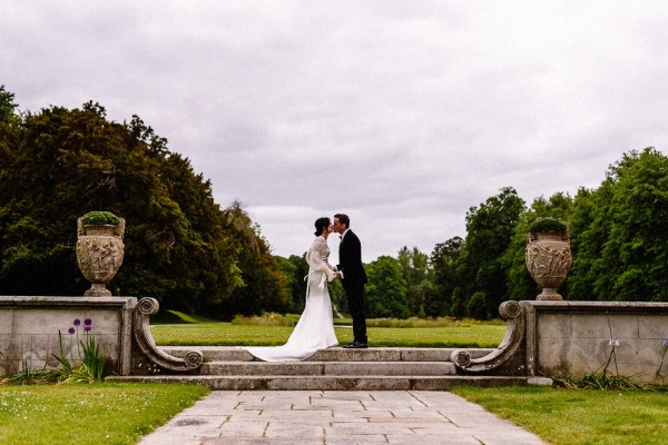 Bride approaches groom in garden grass in background they embrace on steps hug