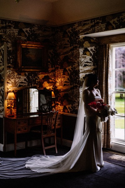 Long train detail on brides gown/dress as she looks out of mirror holding bouquet