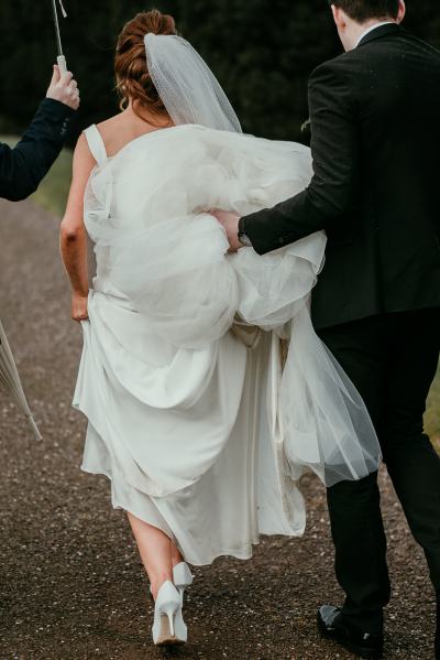 Groom helps bride with bridal gown dress train along pathway